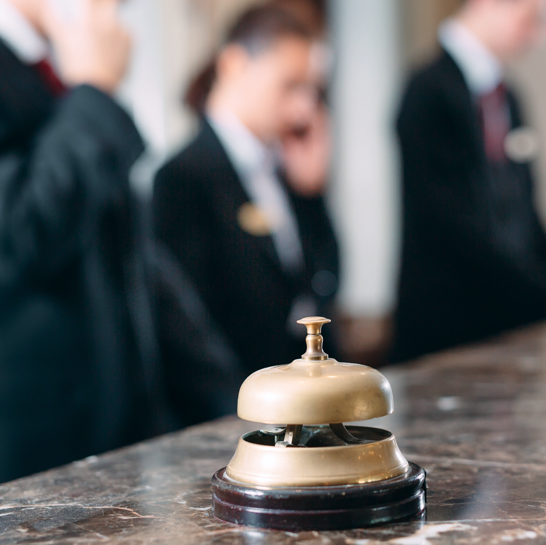 In focus closeup of a hotel front desk bell on a marble counter with blurred employees answering phones in the background