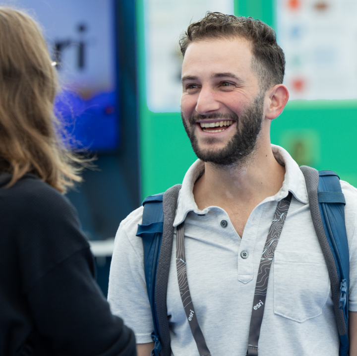 Smiling, bearded attendee with a lanyard and backpack interacts with a presenter at a conference