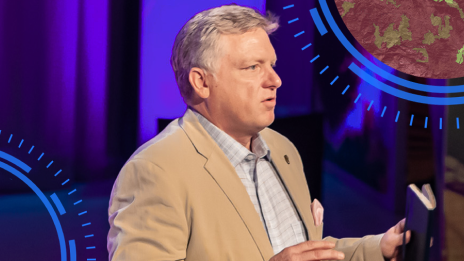 Presenter in a tan jacket and checked shirt gestures with both hands while speaking onstage