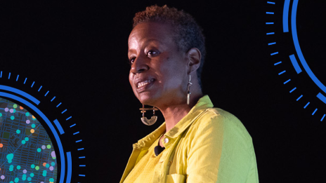 Dark-skinned presenter in a bright yellow jacket speaking onstage