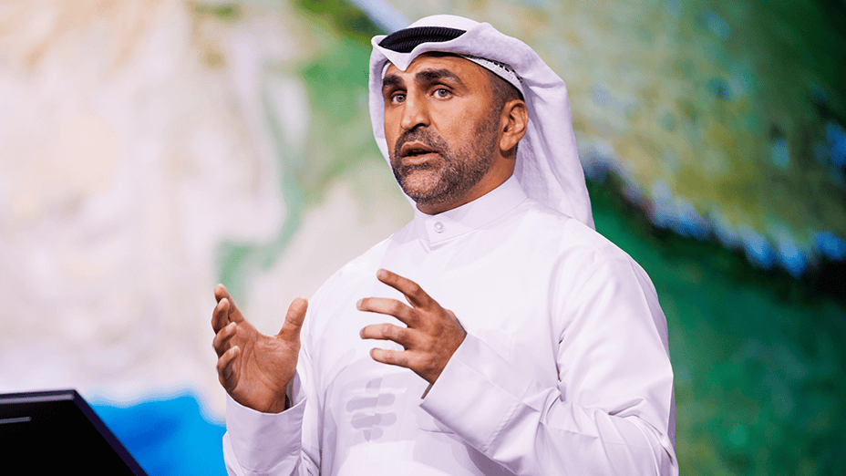 A presenter in a white thawb and ghutra gestures with open hands as they speak in front of a blurred colorful background