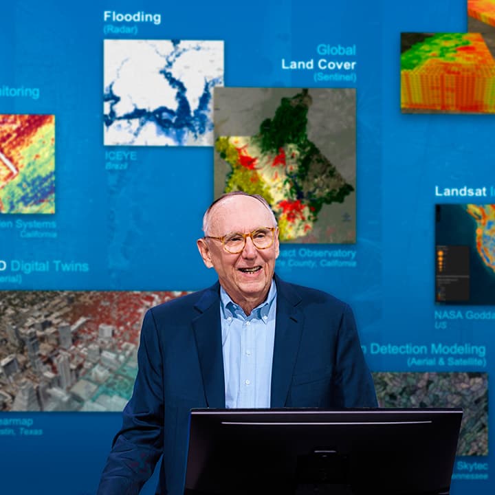 Esri president Jack Dangermond speaking onstage, with an array of colorful maps on a blue background behind him