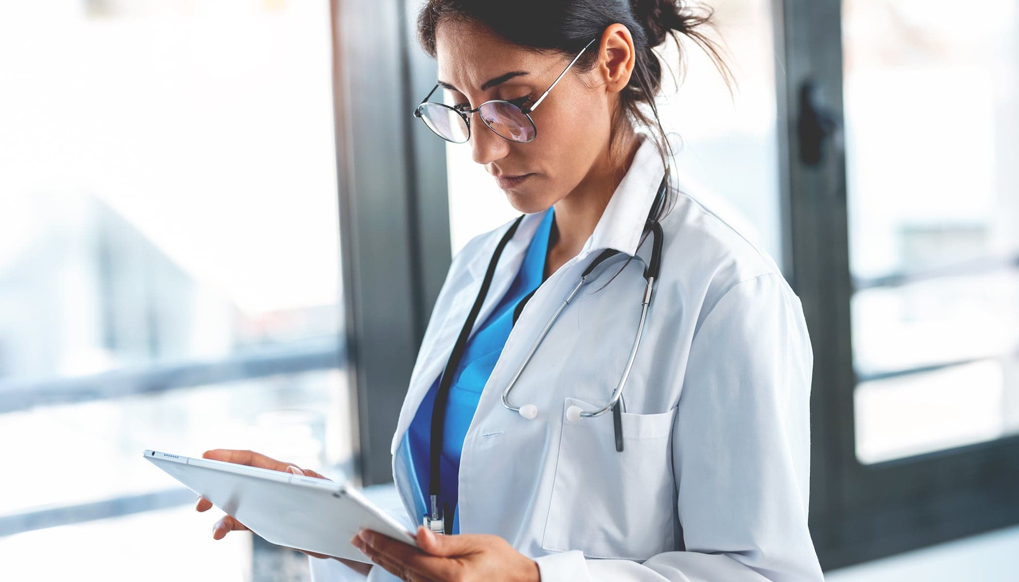 A doctor wearing blue scrubs and a white lab coat reading a handheld tablet beside a wall of bright windows