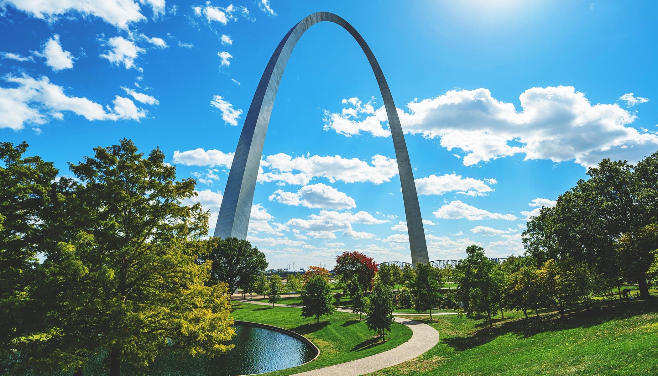 The St. Louis Gateway Arch with a bright green tree-filled park in the foreground and a vivid blue cloud-swept sky overhead