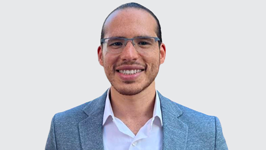 Portrait of Hesham smiling in a white collared shirt and light blue blazer with a plain white background