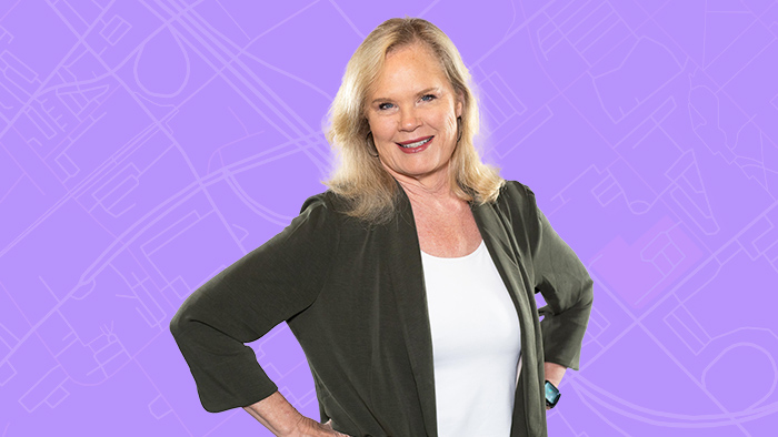 A smiling person wearing a white blouse and dark blazer posing with hands on hips with a flat violet background