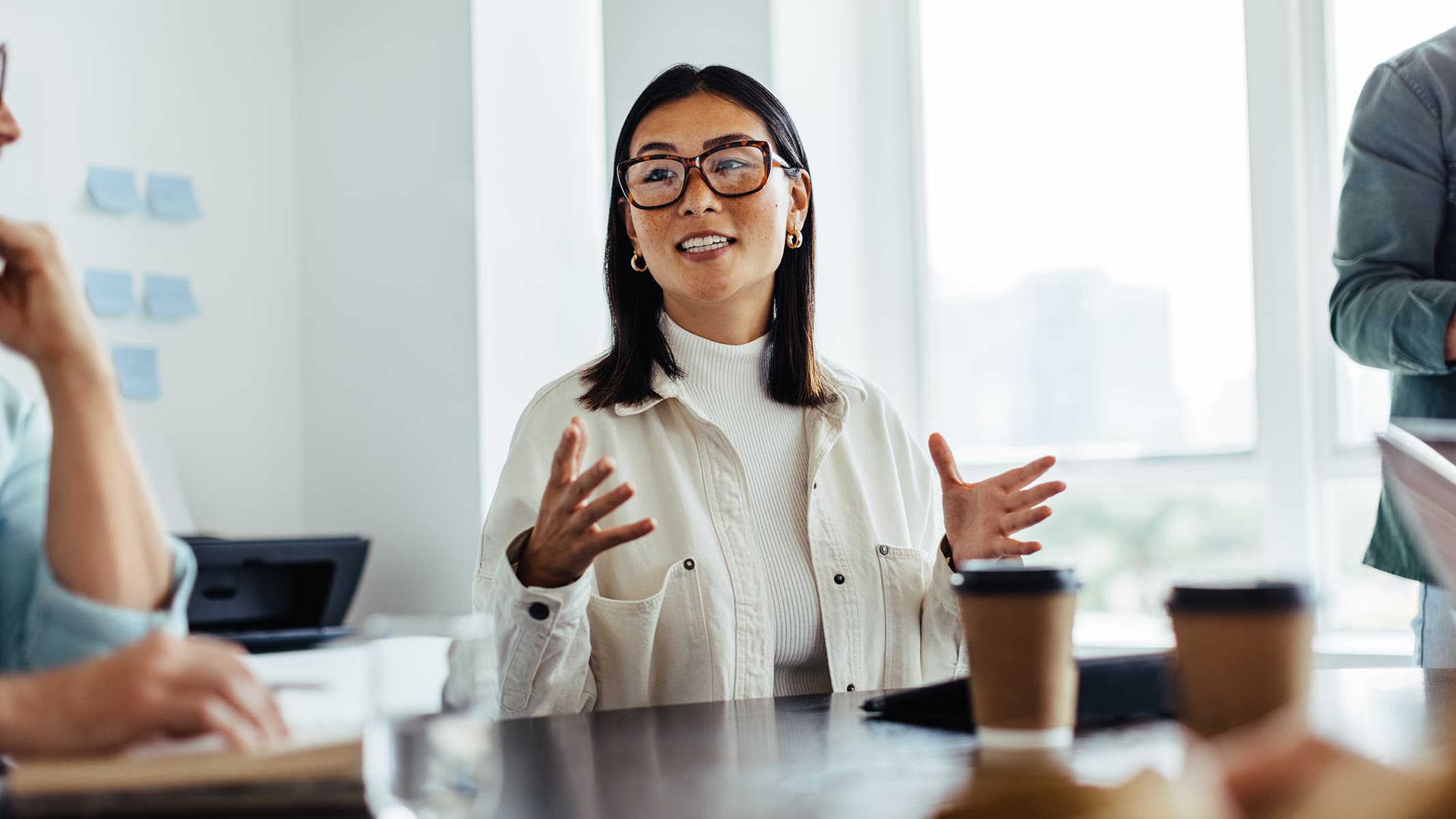 A business professional wearing a white high-necked sweater gesturing with her hands as she explains something with several colleagues at a conference table