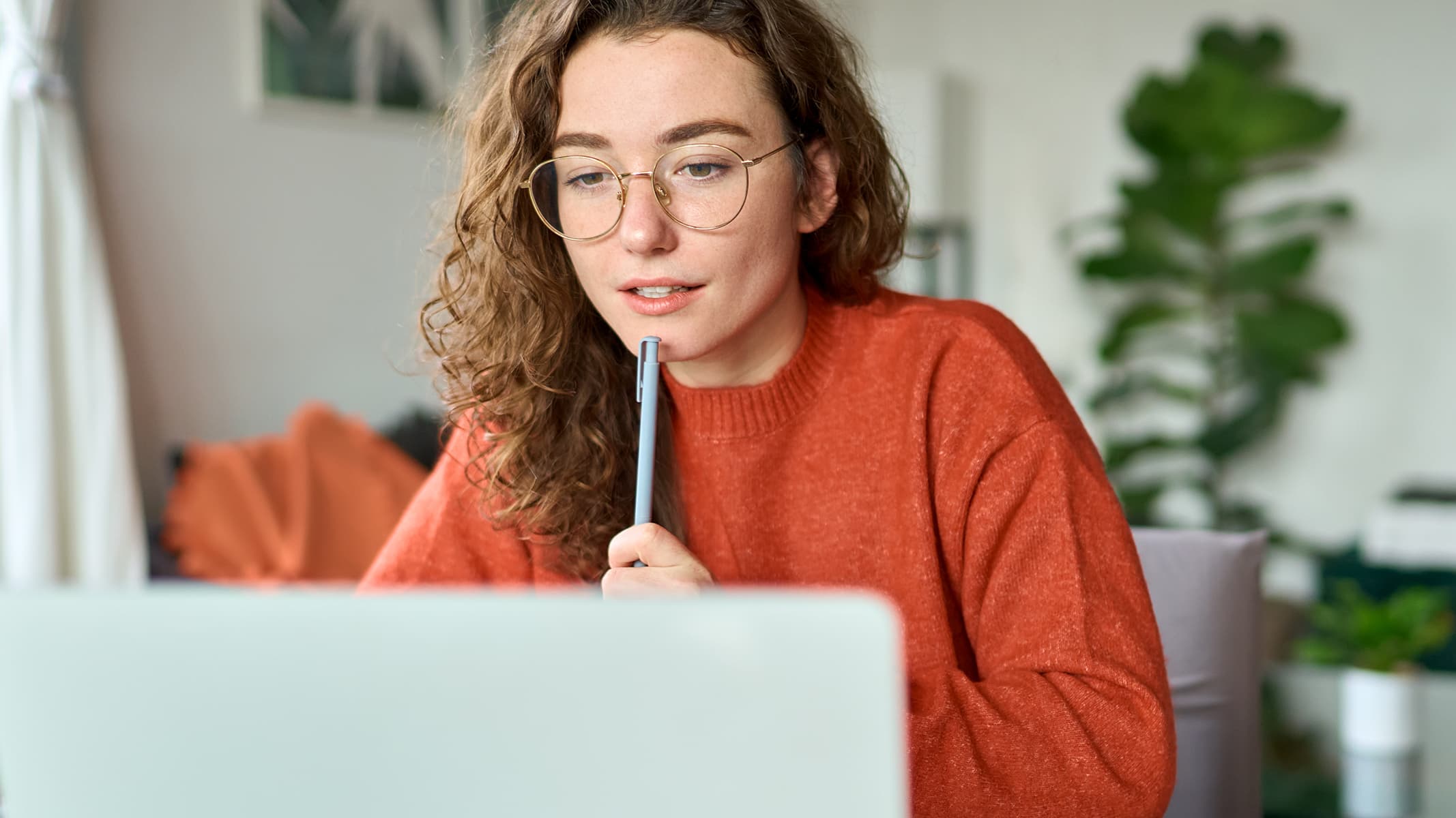 A person wearing an orange sweater gazing contemplatively at a laptop display in a sunlit home office