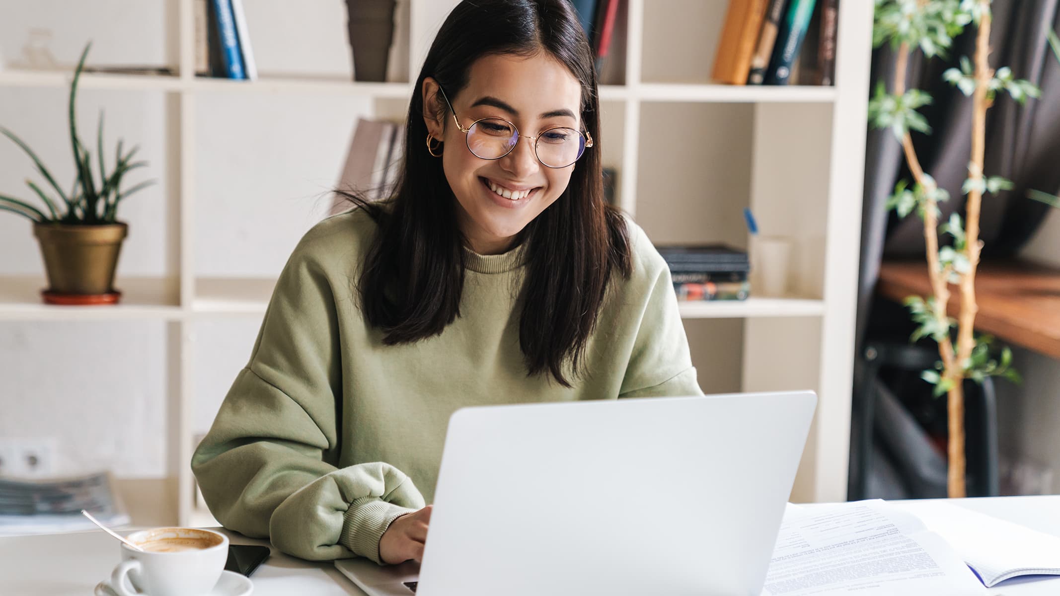 A smiling person wearing a pale green sweater using a laptop in a sunlit home office