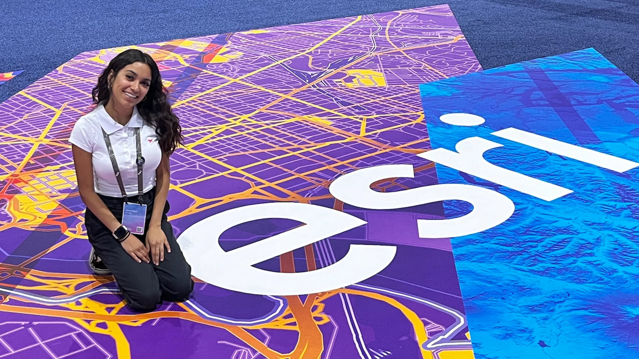 A business professional wearing a conference lanyard posing beside a large Esri logo printed on a conference hall floor