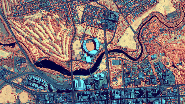 Aerial view of a city with a large stadium in the center, shown in a virtual color palette with blue, red and orange colors  