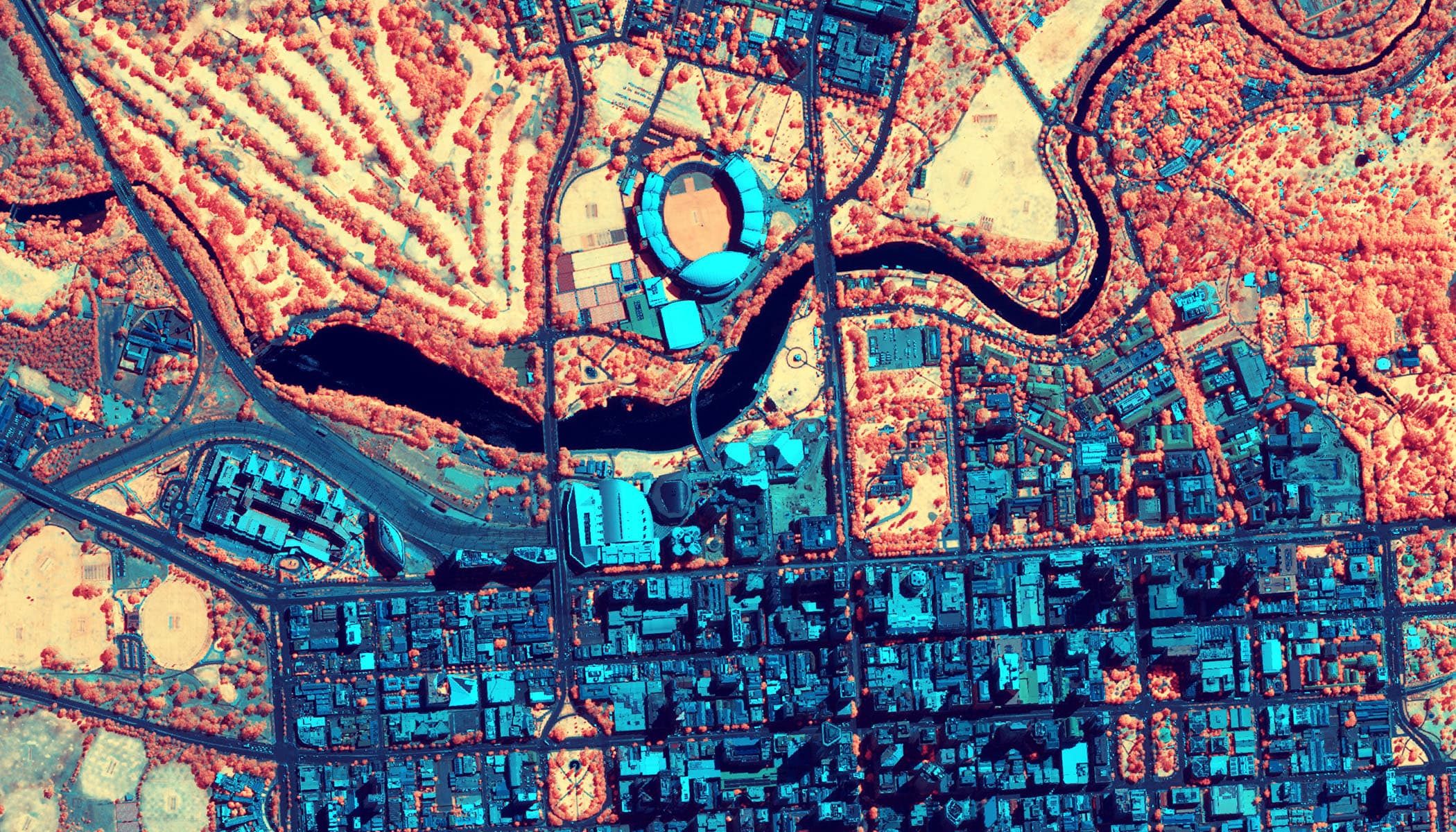 Aerial view of a city with a large stadium in the center, shown in a virtual color palette with blue, red and orange colors  