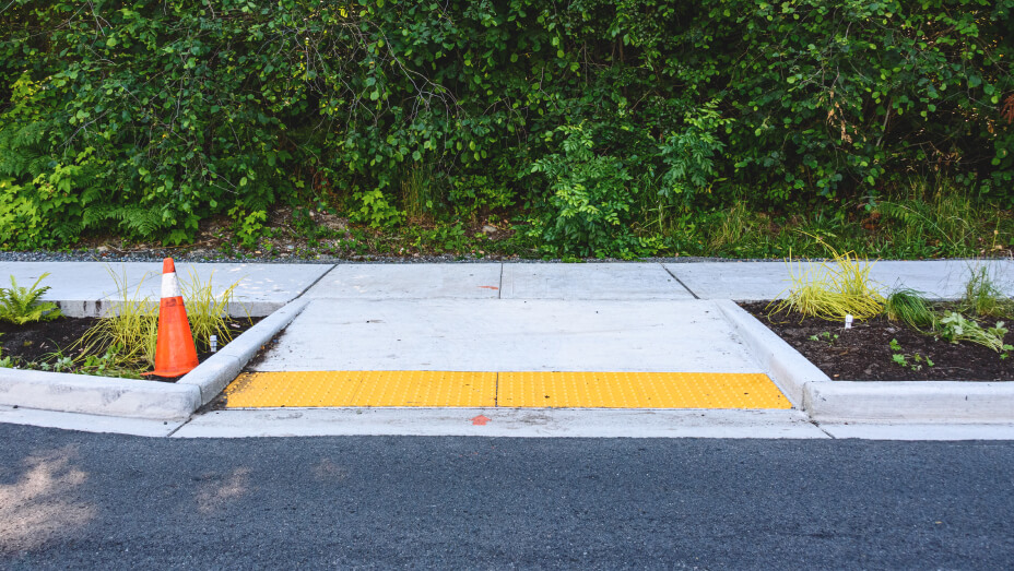 An ADA-compliant curb ramp leading to a sidewalk lined with green foliage