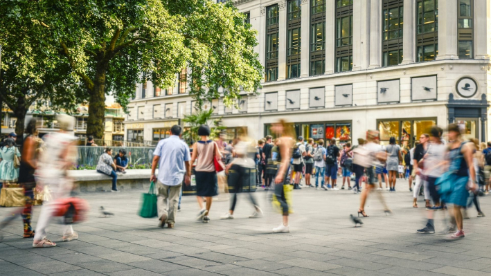 Sonnenbeschienener Platz in einer Einkaufsstraße mit zahlreichen vorbeigehenden Fußgängern, die leger gekleidet sind