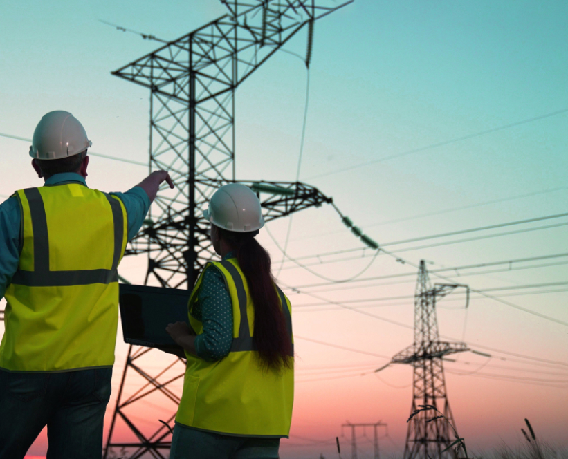 Two workers in yellow vests and hard hats pointing at power lines outdoors and holding a computer