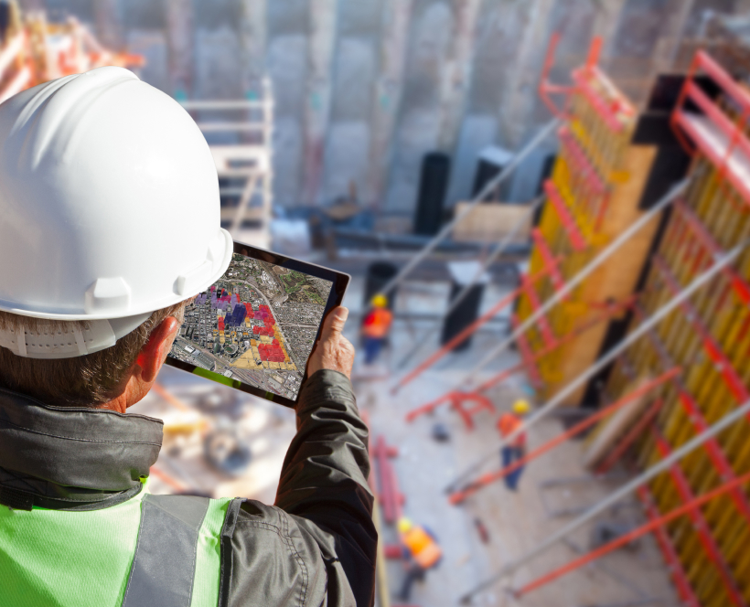 Worker at construction site in a hard hat, looking at iPad showing 3D city model with red and purple highlighted buildings
