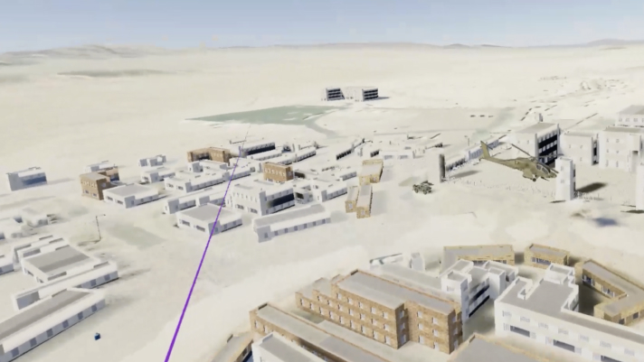 Rappresentazione virtuale di una base nel deserto con edifici 3D grigi e beige, un elicottero e una linea viola 