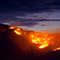 A panorama image of large sloping hillsides covered by a brightly glowing wildfire beneath a dark purple smoke-filled twilight sky