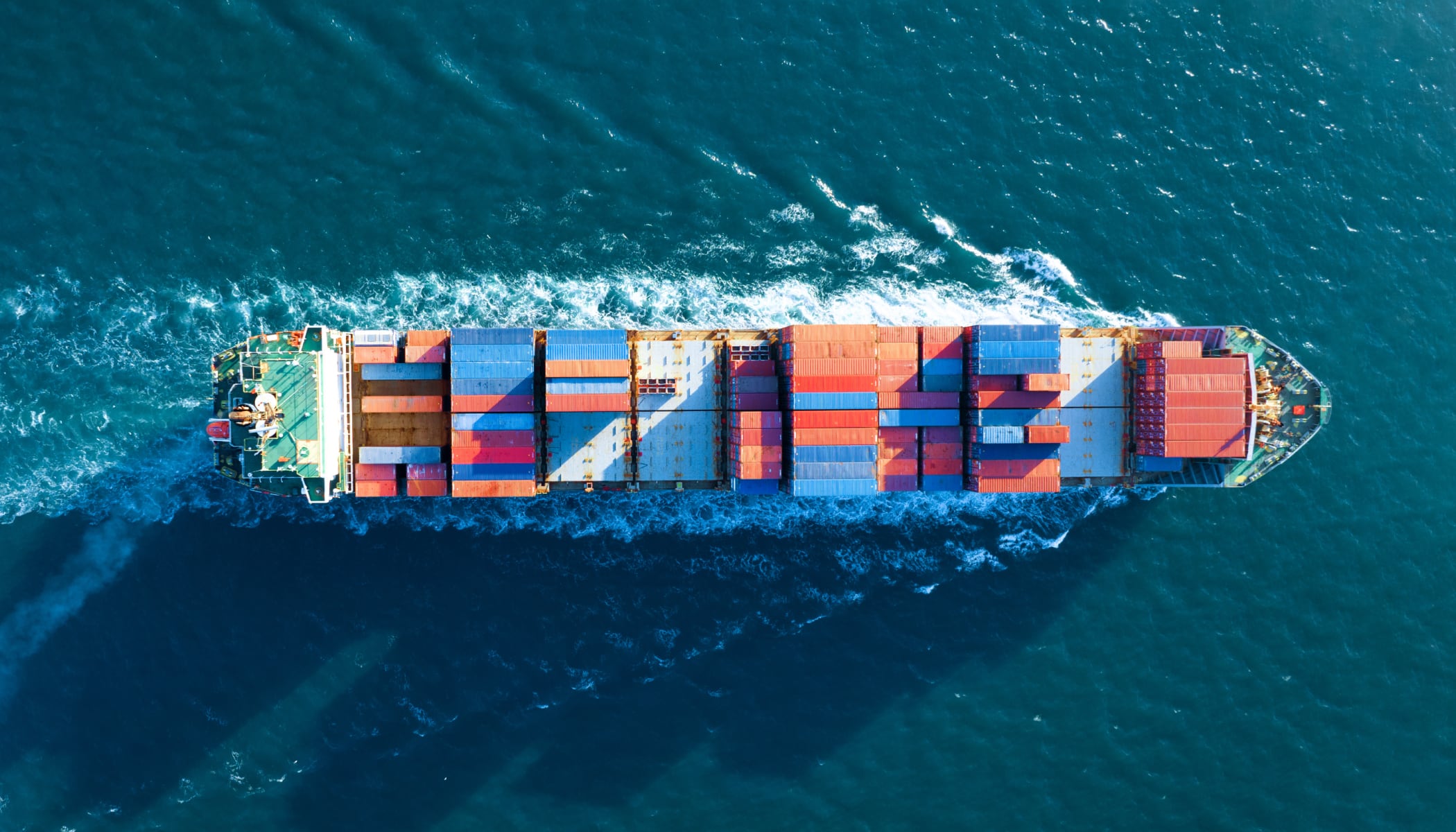 Aerial view of a cargo ship loaded with rows of colorful cargo containers sailing through rippling green waters