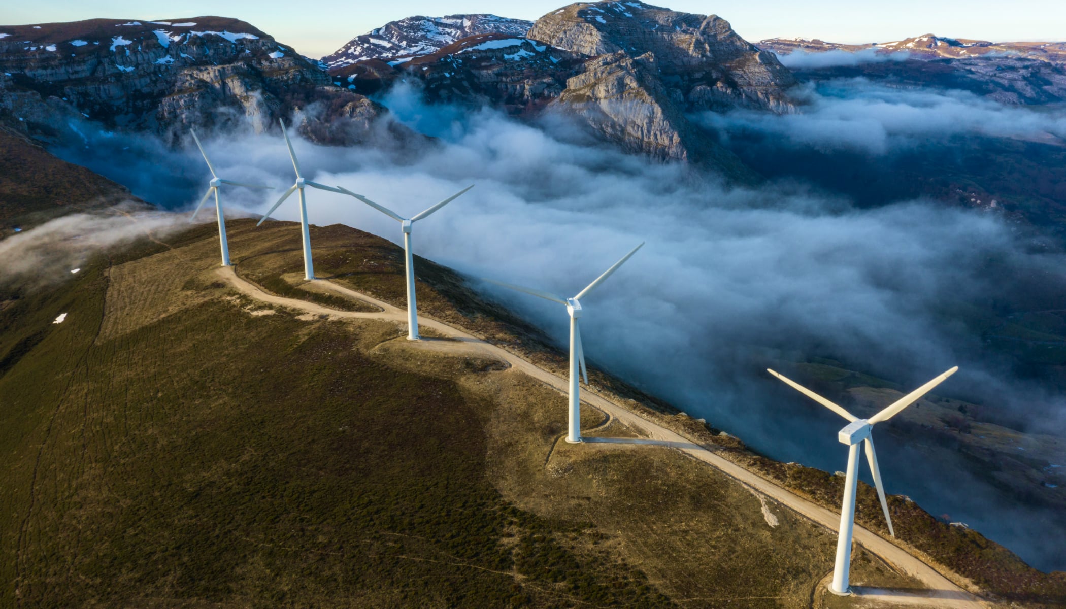 A row of white wind turbines along the crest of a green hilltop surrounded by a layer of fog through which rolling hillsides are visible