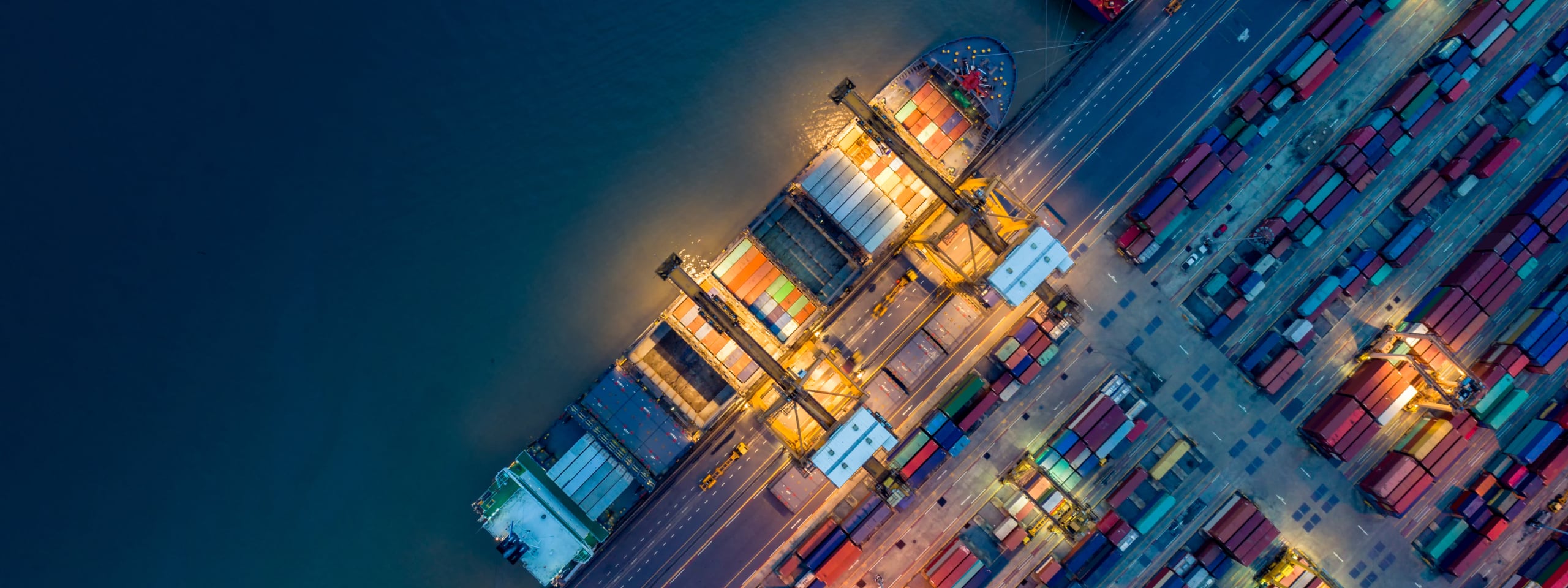 Aerial view of a container ship at port in rippling dark blue waters, docked at a shipping yard warmly lit for nighttime with many colorful cargo containers 