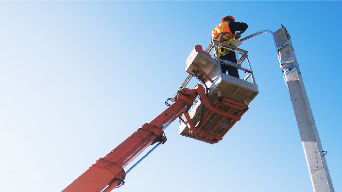 A person wearing an orange safety vest standing on the platform of an extended utility crane to reach the top of a pole