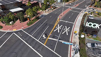 An aerial view of a four-lane, two-way street near an intersection surrounded by buildings