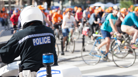 Duża grupa rowerzystów w pomarańczowych kaskach na oświetlonej słońcem ulicy z policjantem na motocyklu na pierwszym planie