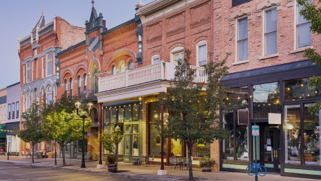 A clean tree-lined city street with brightly lit retail storefronts under a pale blue sky