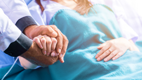 A person wearing a lab coat holding the hand of a patient in a hospital bed under a blue blanket