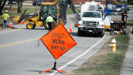 Several utility and construction vehicles on a city street with an orange sign in the foreground stating “Utility Work Ahead”