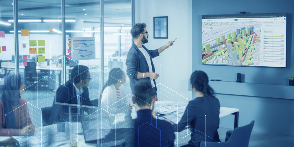 A man presenting a 3D city model on a television screen in front of people in a conference room