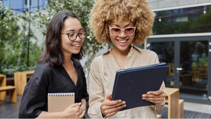 Duas mulheres sorrindo enquanto olham para um tablet do lado de fora de um prédio 