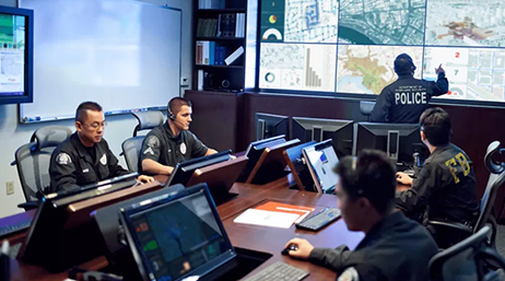 A group of police offers using computers at a conference table in front of a wall of multiple screens displaying maps  