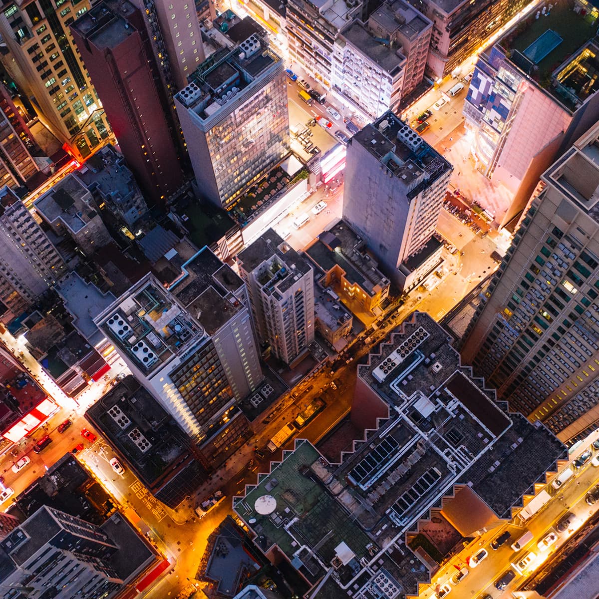 An aerial view of a dense city at night, with large and tall buildings and streetlights illuminating the streets below