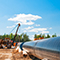 A large pipeline resting on the ground near workers and heavy machinery under a bright blue sky