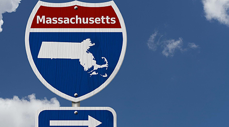 Blue and red road sign that says “Massachusetts” across the top with the state’s silhouette at the bottom, with a cloudswept blue sky in the background