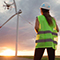 A utility worker wearing yellow safety gear stands facing a cloudy golden sunset 