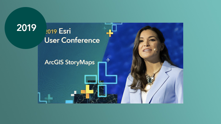 A person in a pale blue suit wearing a headset speaking onstage against a deep blue background overlaid with geometric green and gold shapes and text stating “2019 Esri User Conference- ArcGIS StoryMaps”