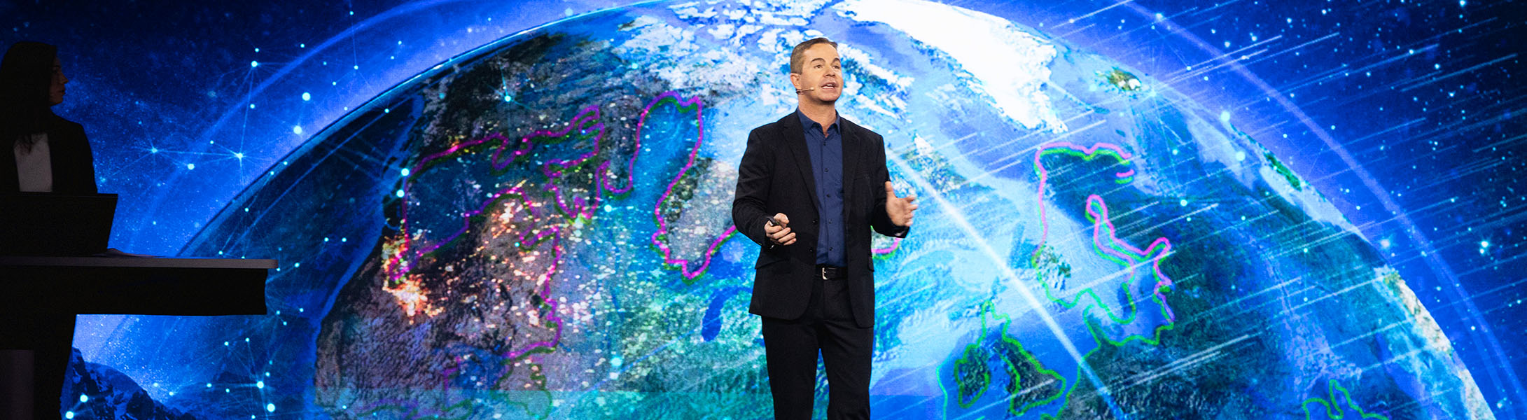 Apresentador falando em um evento da Esri tendo como pano de fundo um globo digital colorido