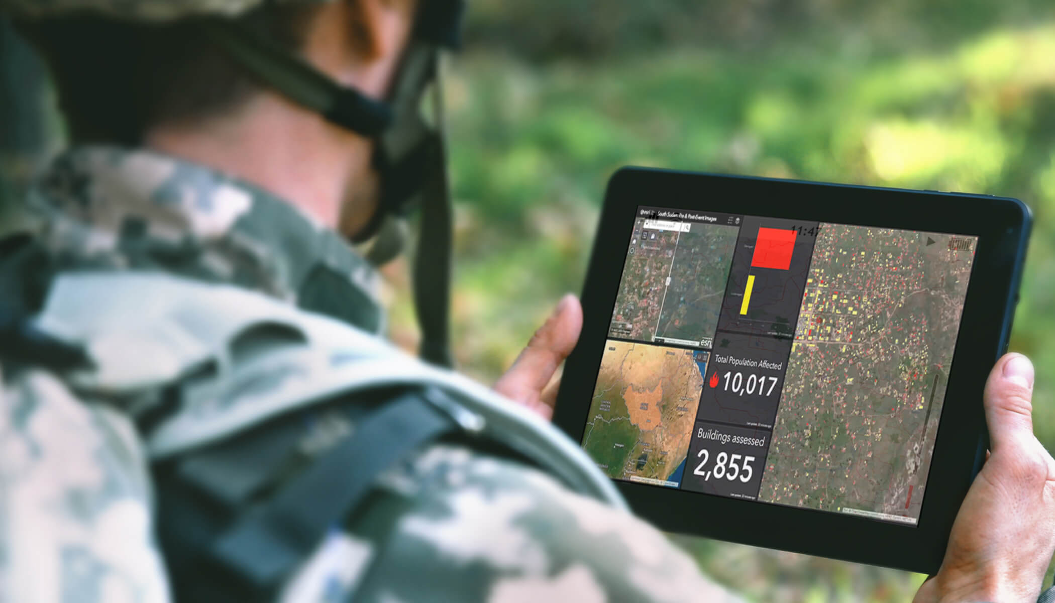 A soldier wearing a combat uniform and helmet holding a tablet displaying a map dashboard