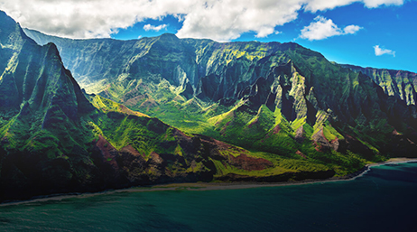 Aerial view of soaring green mountains meeting the shoreline beneath a bright blue sky