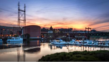 在橙色和深蓝色的夕阳映照下，一座商业建筑群在静止的码头旁点亮了夜灯，码头上停满了白色的船只