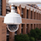 A close-up view of a white security camera mounted on a pole with a large brick tree-lined building in the background