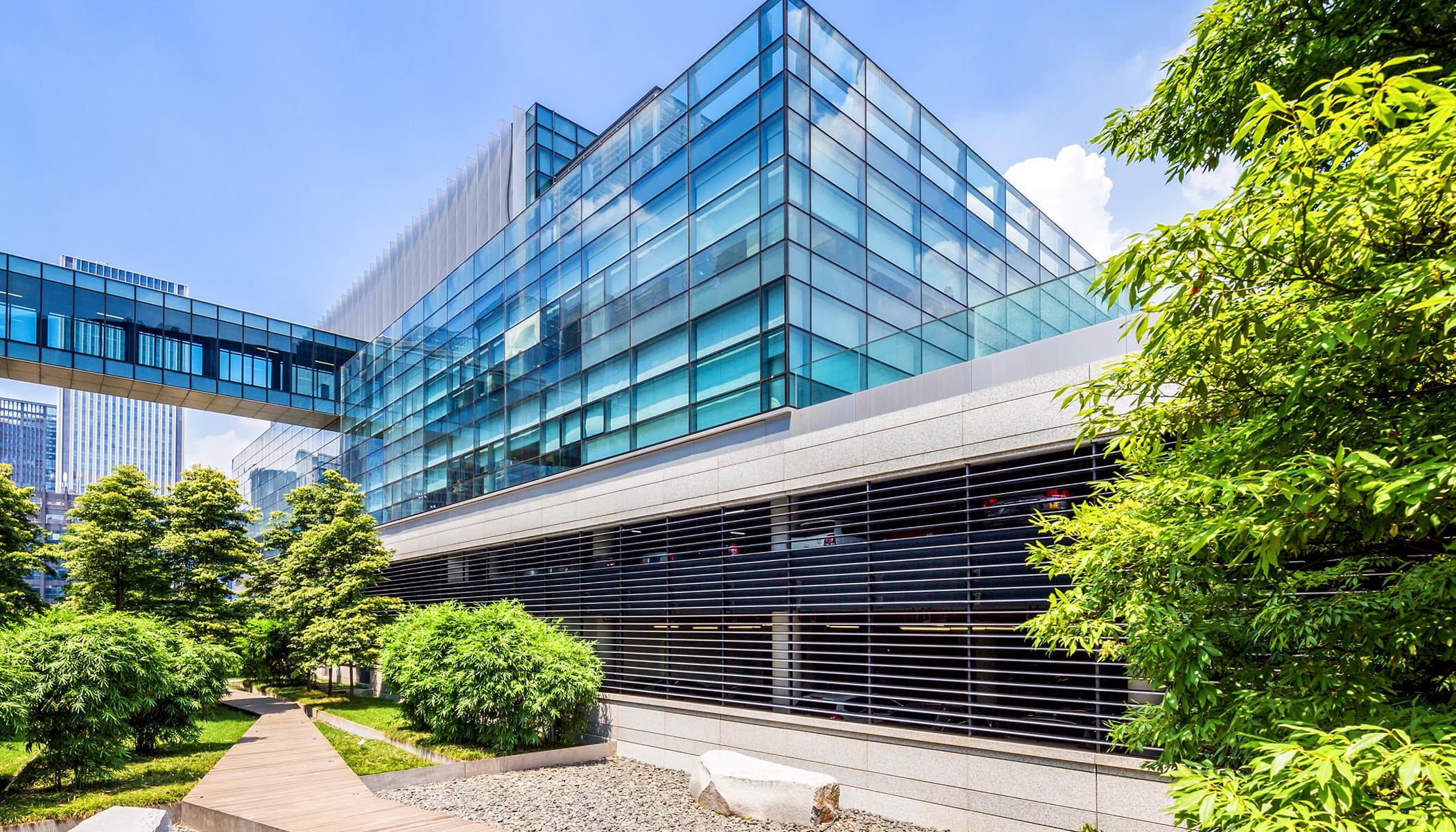 Modern office building with a glass facade and tree-lined pedestrian bridge, under a clear blue sky with white clouds.