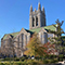 A historic college building of stone topped with a clock tower surrounded by autumnal trees under a clear blue sky