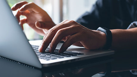 Фотография крупным планом пары рук с часами, набирающих текст на клавиатуре ноутбука в домашнем офисе