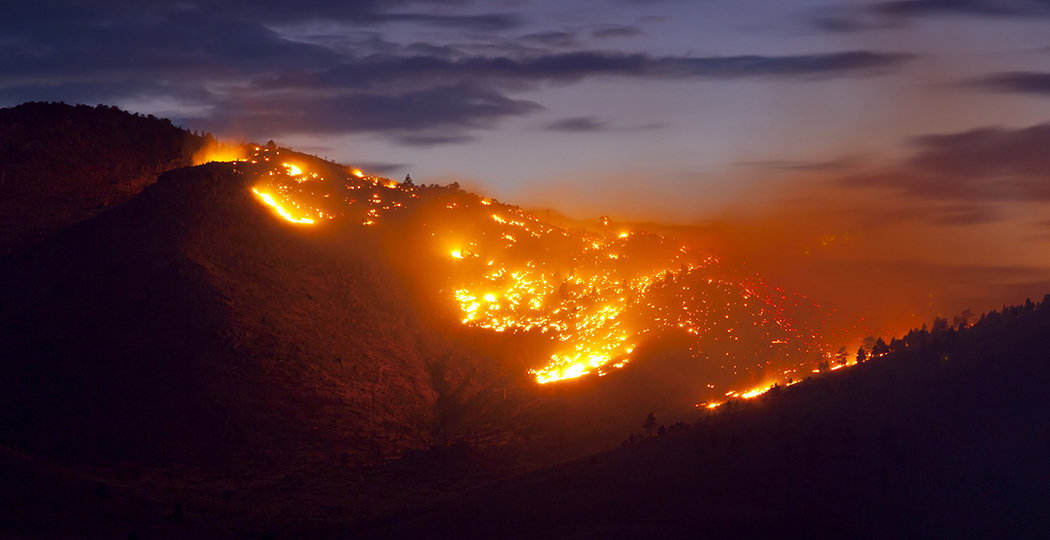 Aerial image of a wildland hillside glowing orange with widespread wildfire as dark smoke drifts into a purple twilight sky