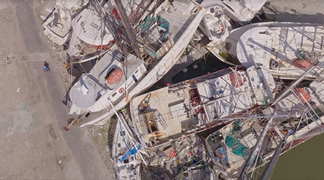 Aerial view of six personal sailboats heaped together in a pile amid waterside post-hurricane rubble