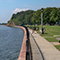 A lush green park with children playing on a fenced sidewalk curving along the edge of a dark blue lake under a pale blue sky